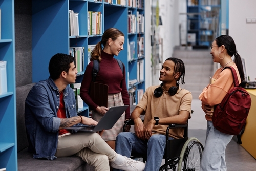 Diverse group of students with young man in wheelchair chatting cheerfully in college library